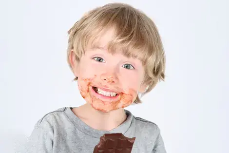 Young child holding unwrapped chocolate bar looking at camera with chocolate residue around mouth.