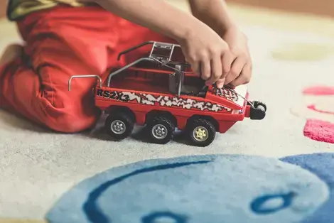 Child playing with red and black vehicle toy on light-colored carpet.