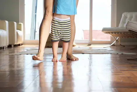 Adult standing behind child on tiled floor. The adult is seen from the thighs down. The child from the waist down.